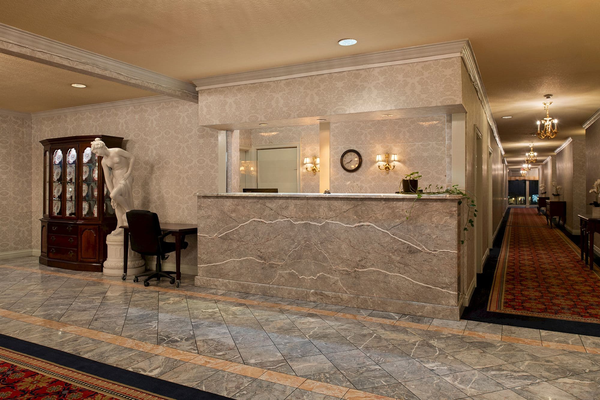 Marble front desk in the lobby, beside a classical statue, a china cabinet and a chandelier-lit corridor