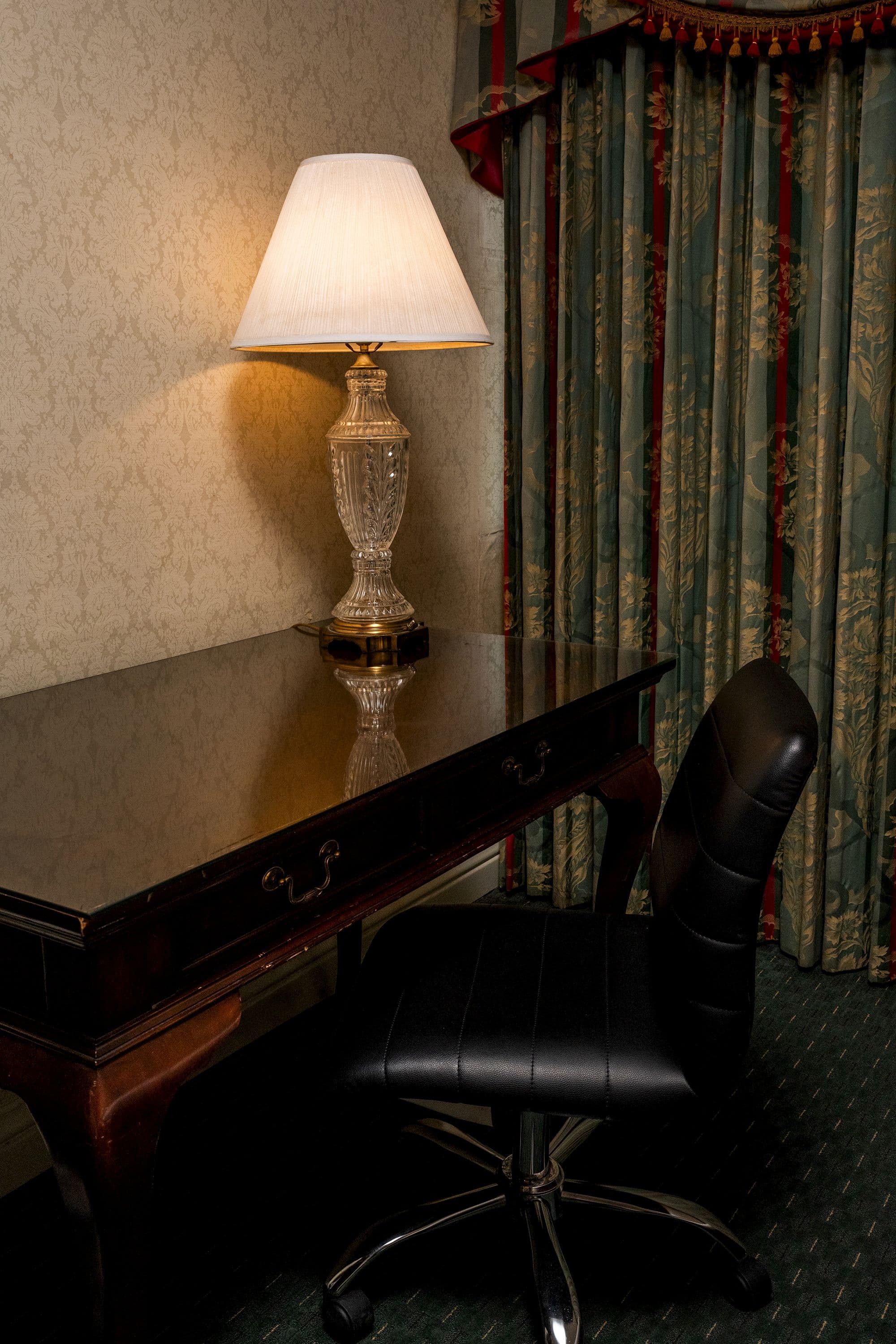Cherry wood writing desk with a crystal lamp and a leather chair beside patterned curtains